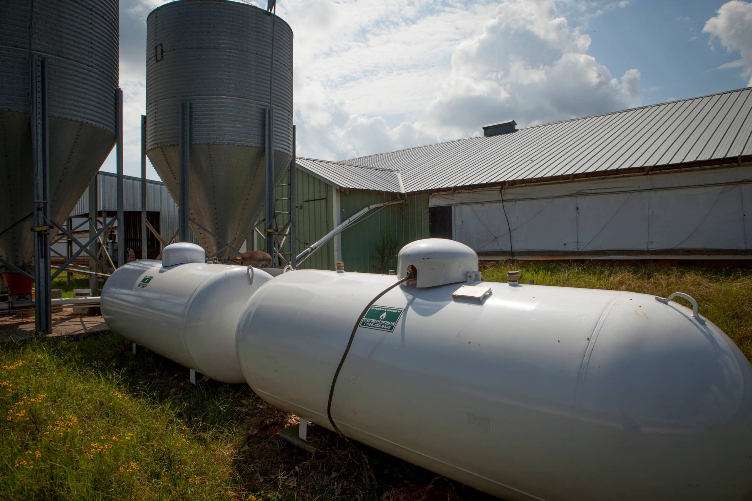 Large white propane tanks rest on grassy ground beside a metal building, with two tall grain silos in the background beneath a sky scattered with clouds.