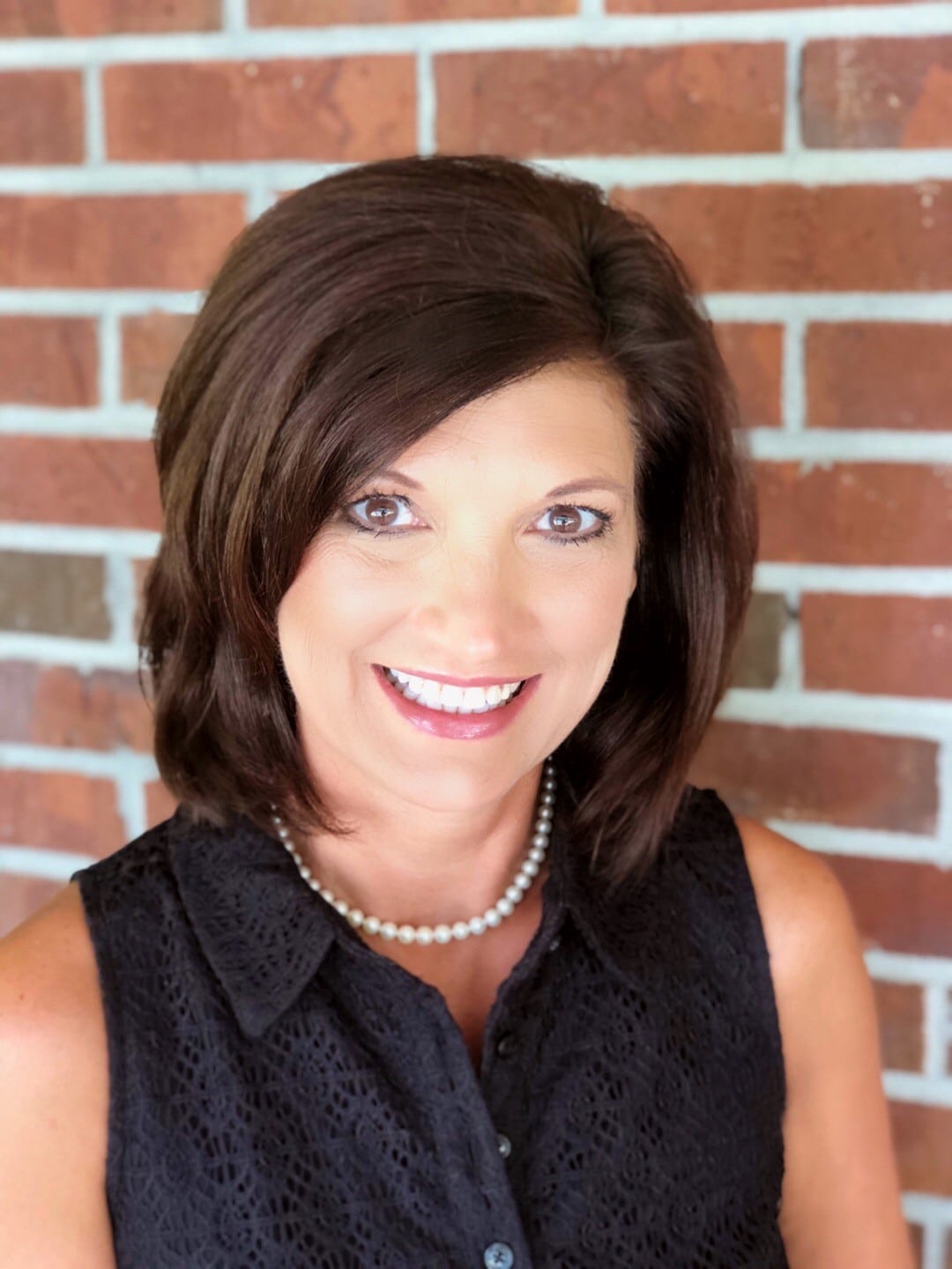 A cheerful woman with short brown hair, dressed in a black sleeveless top and pearl necklace, stands smiling in front of a brick wall.
