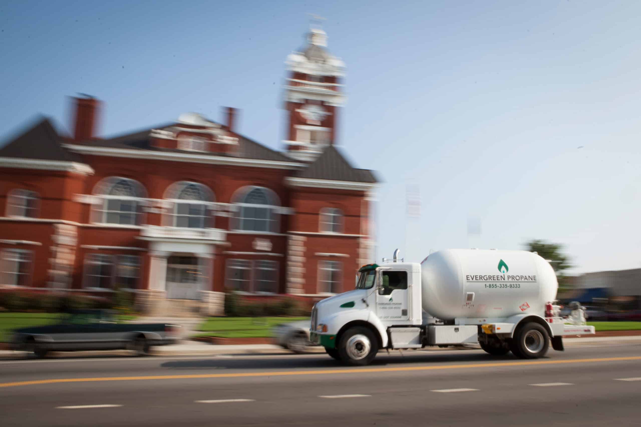 A white Evergreen Propane truck is driving by a red brick building with a clock tower on a sunny day, with the blurred background showing the motion of the truck.