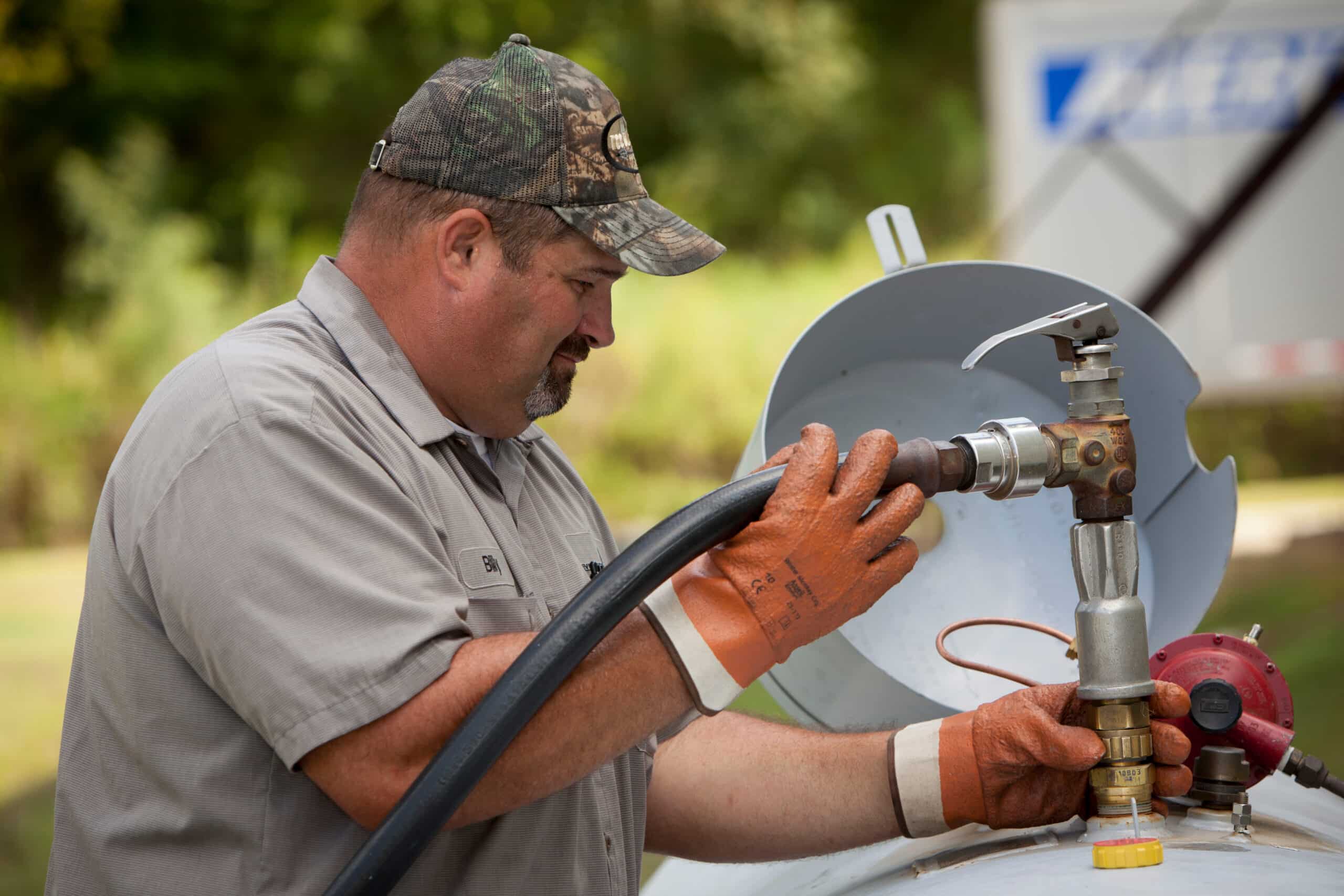 A man wearing work gloves and a camouflage hat attaches a hose to a propane tank outside, surrounded by green trees.