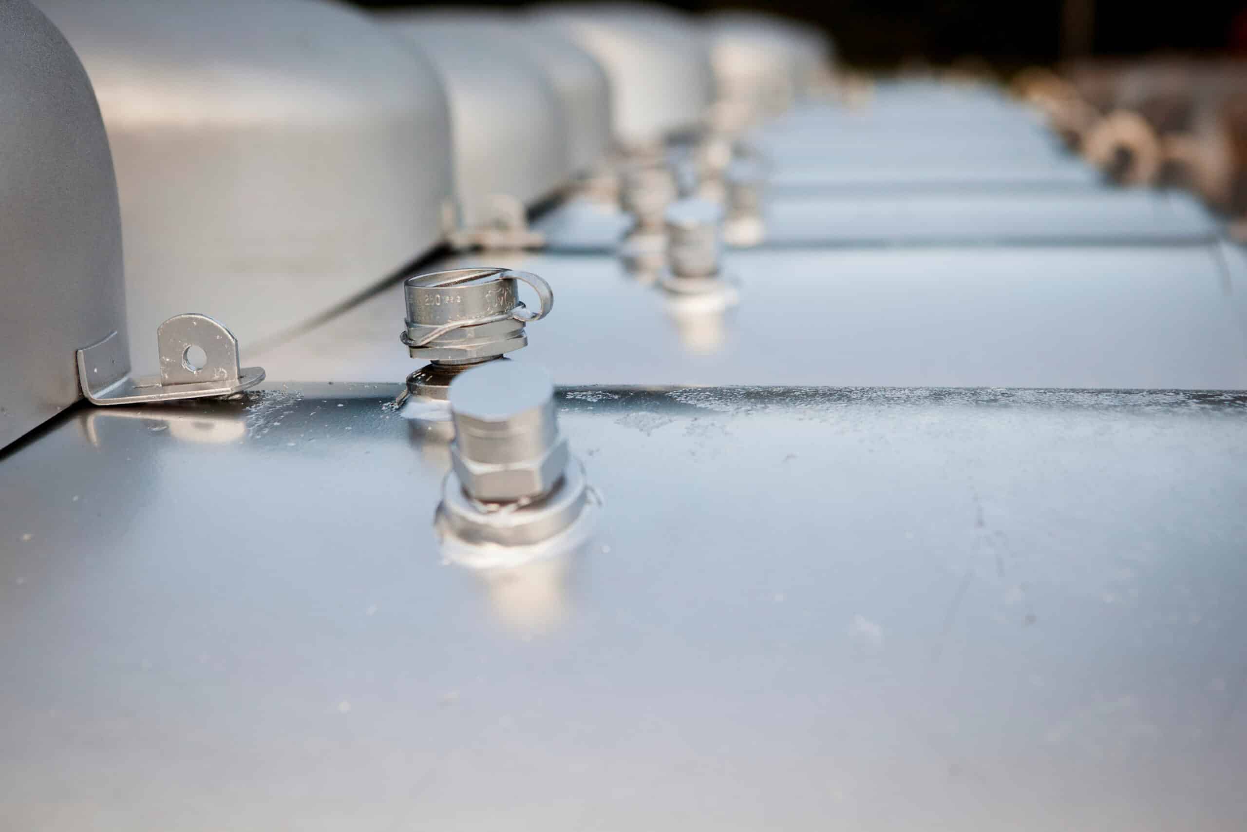 A close-up of shiny metallic tanks or containers with bolts and secure caps, neatly arranged in a row outdoors and reflecting sunlight.