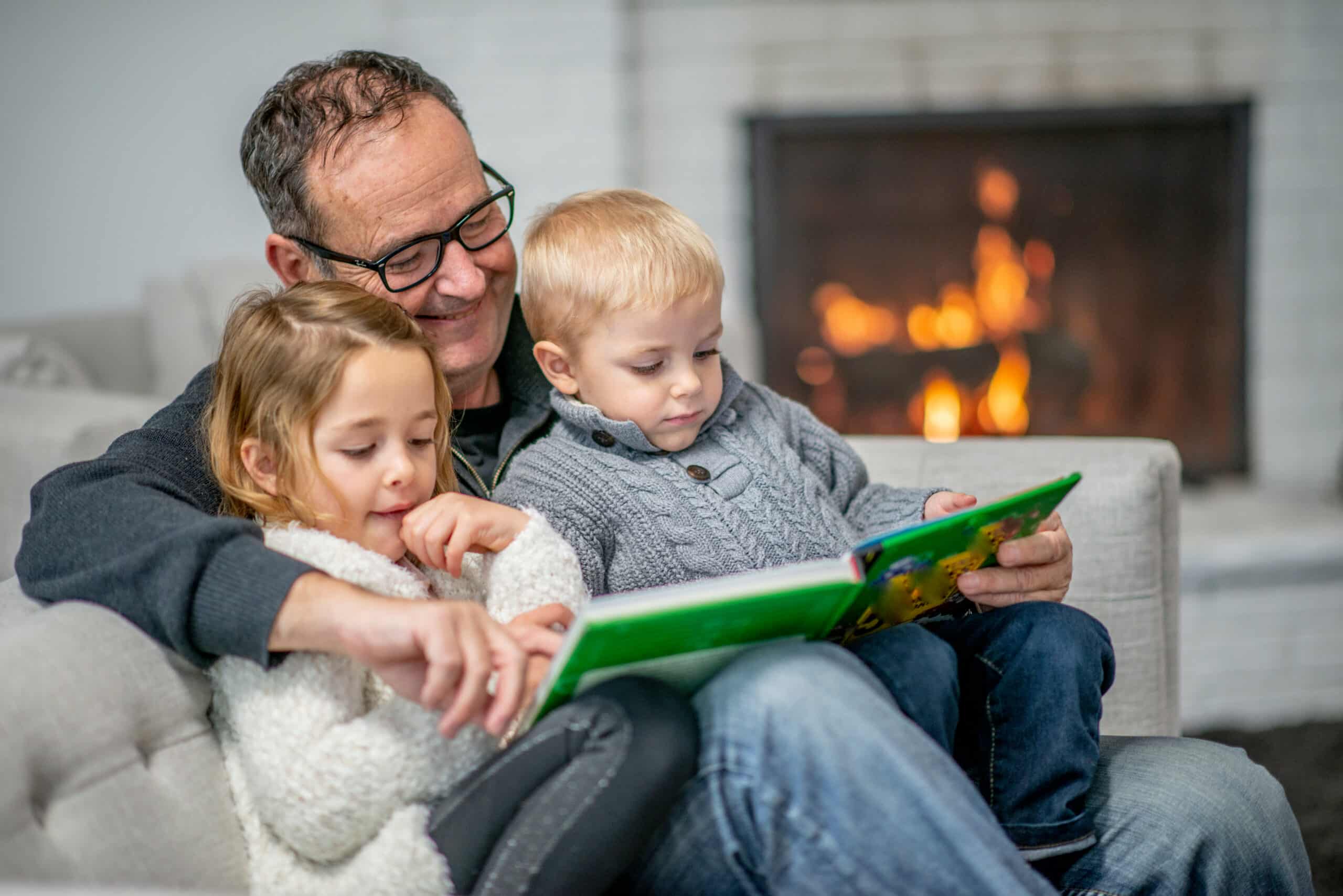 An older man reads a book to a young boy and girl as they sit together on a couch in front of a fireplace. Both children focus on the book and listen attentively.