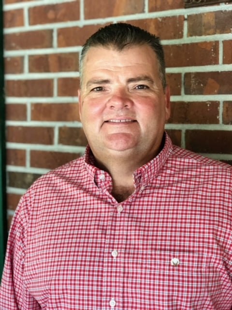 A man in a red and white checkered button-up shirt stands before a brick wall, smiling at the camera.