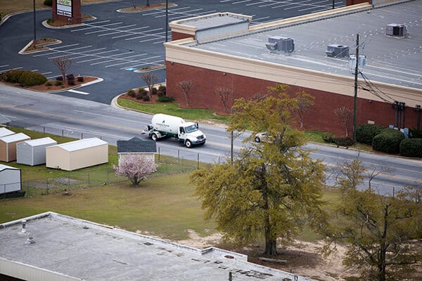 A white utility truck drives along a quiet street next to a large, mostly empty parking lot and a few small storage sheds. Nearby, there are trees and some blooming bushes.