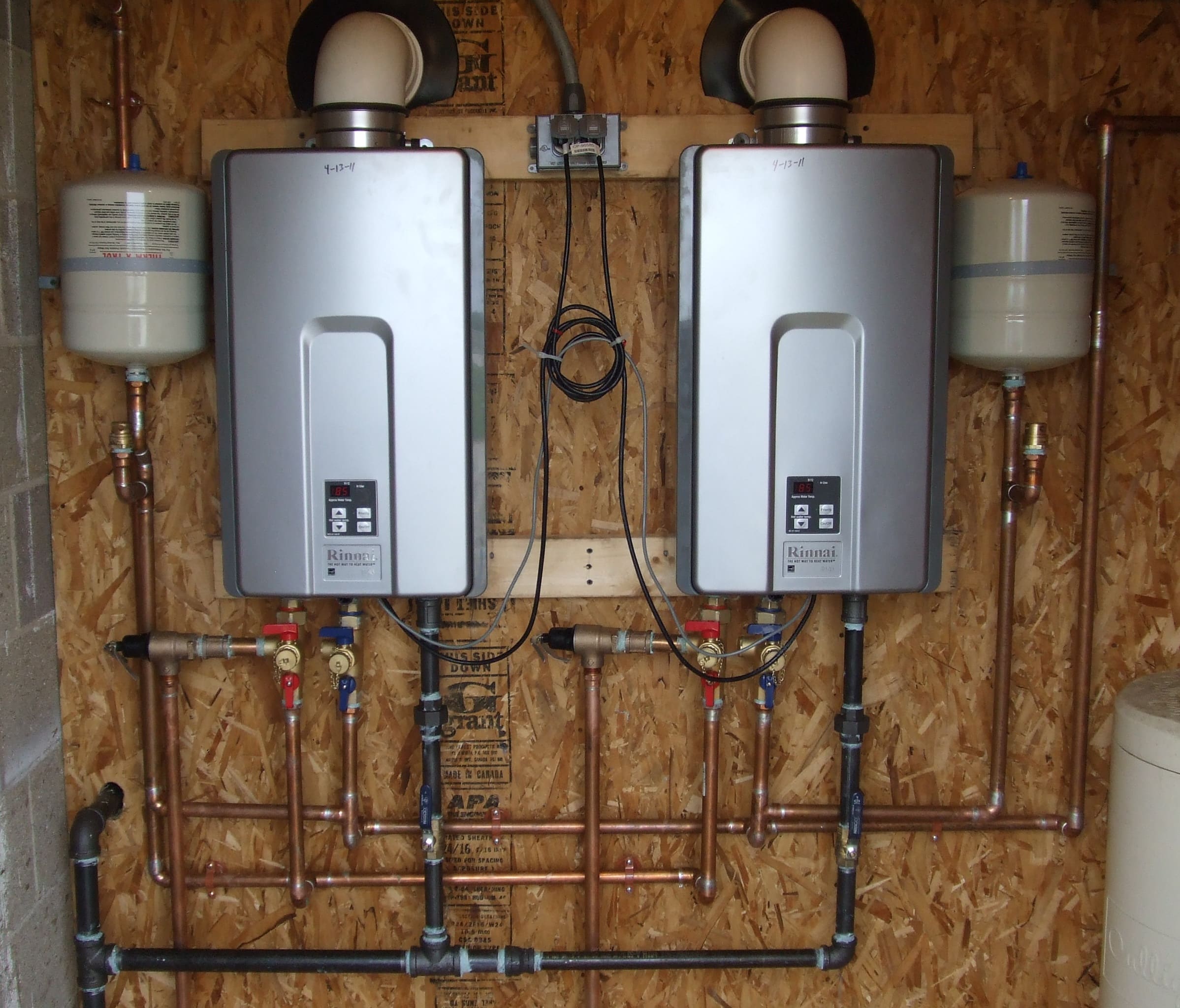 Two tankless water heaters mounted on a plywood wall, each connected to several copper pipes and flanked by two expansion tanks, with visible wiring and pipe fittings underneath.