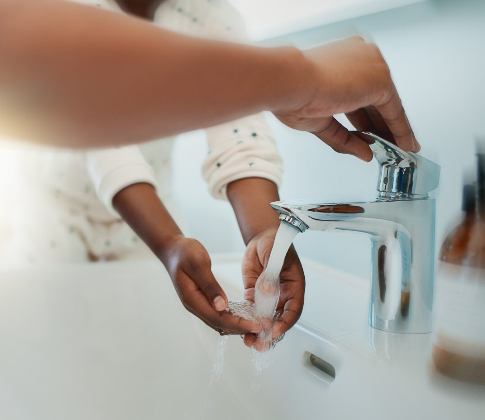 A child washes their hands under running water at a sink while an adult assists by turning on the faucet. The emphasis is on their hands and the flowing water, with bright light illuminating the background.