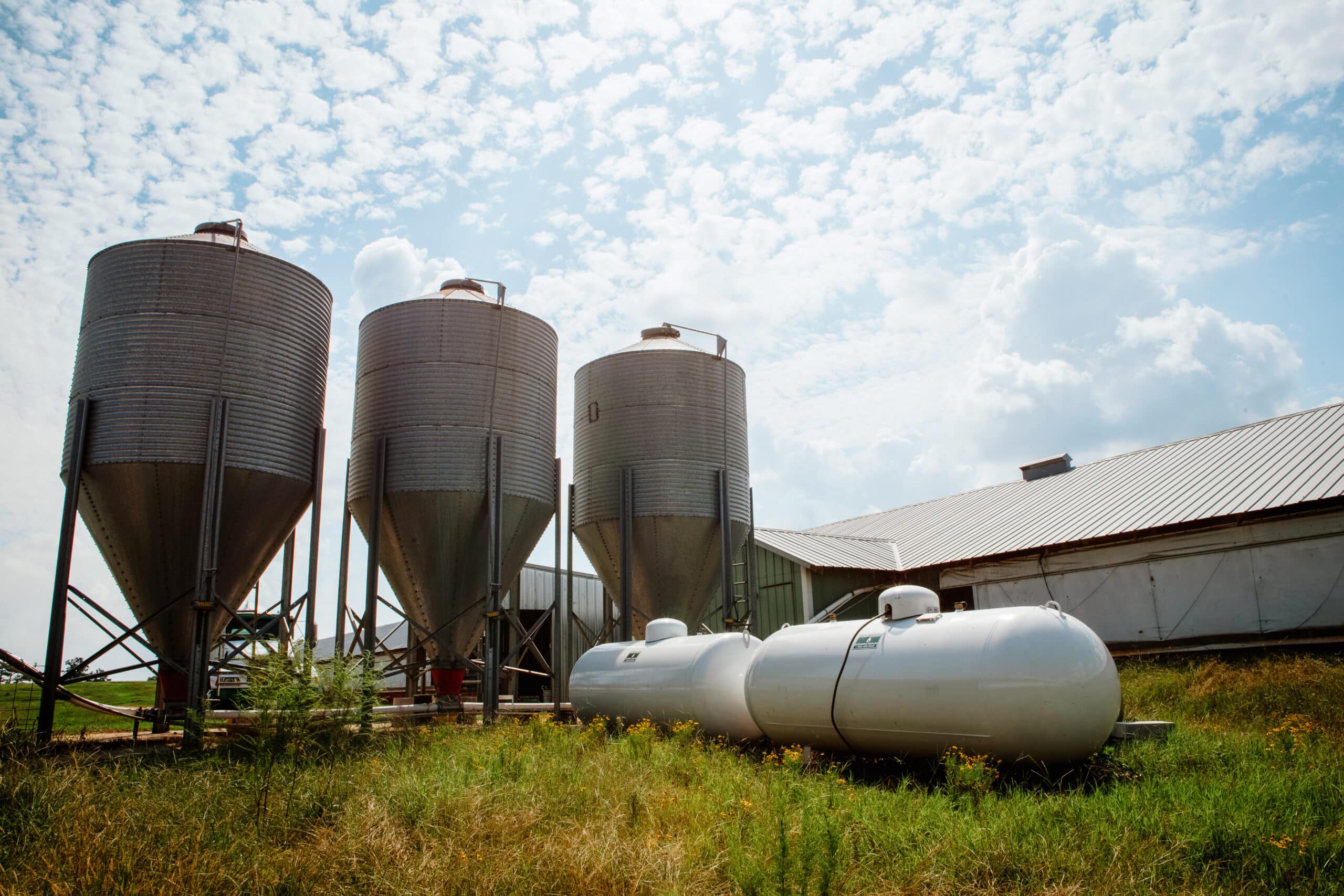 Four metal silos and two large white propane tanks rest on a grassy area beside a barn with a corrugated metal roof, beneath a partly cloudy sky.
