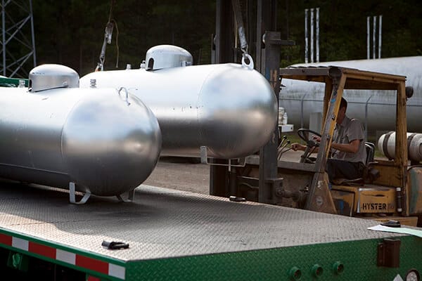 A worker uses a forklift outdoors to load a large silver propane tank onto a truck bed, while several other tanks are visible in the background.