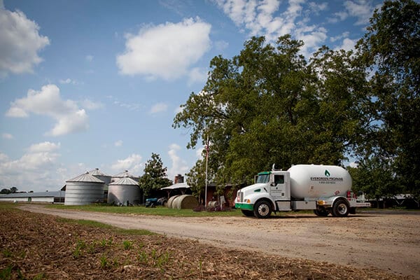 A white propane delivery truck is parked on a dirt road next to tall trees and multiple silver grain silos on a rural farm, all beneath a partly cloudy sky.